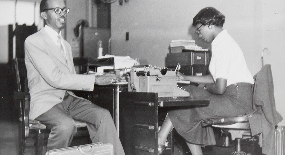 Scheltes collection archival greyscale photograph of a Black blind man with a briefcase, sitting beside a desk. A Black woman is sitting at the desk, looking down and writing.