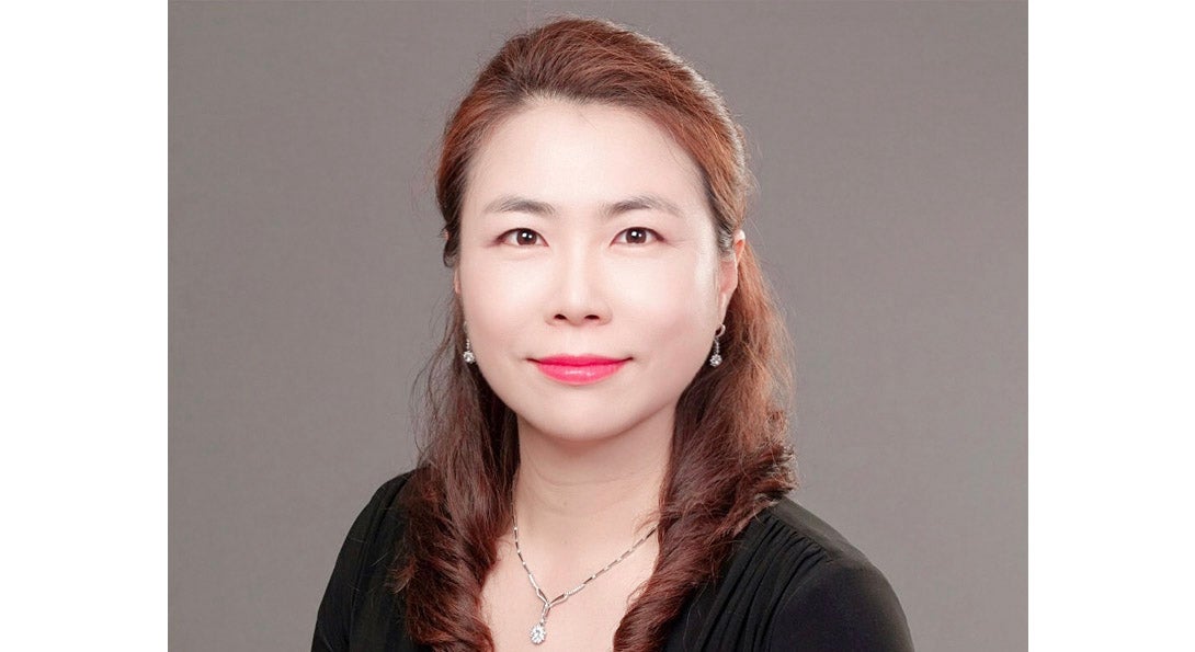 Portrait of Jung Mi Scoulas who is wearing black blouse and necklace and earrings in front of a neutral background.